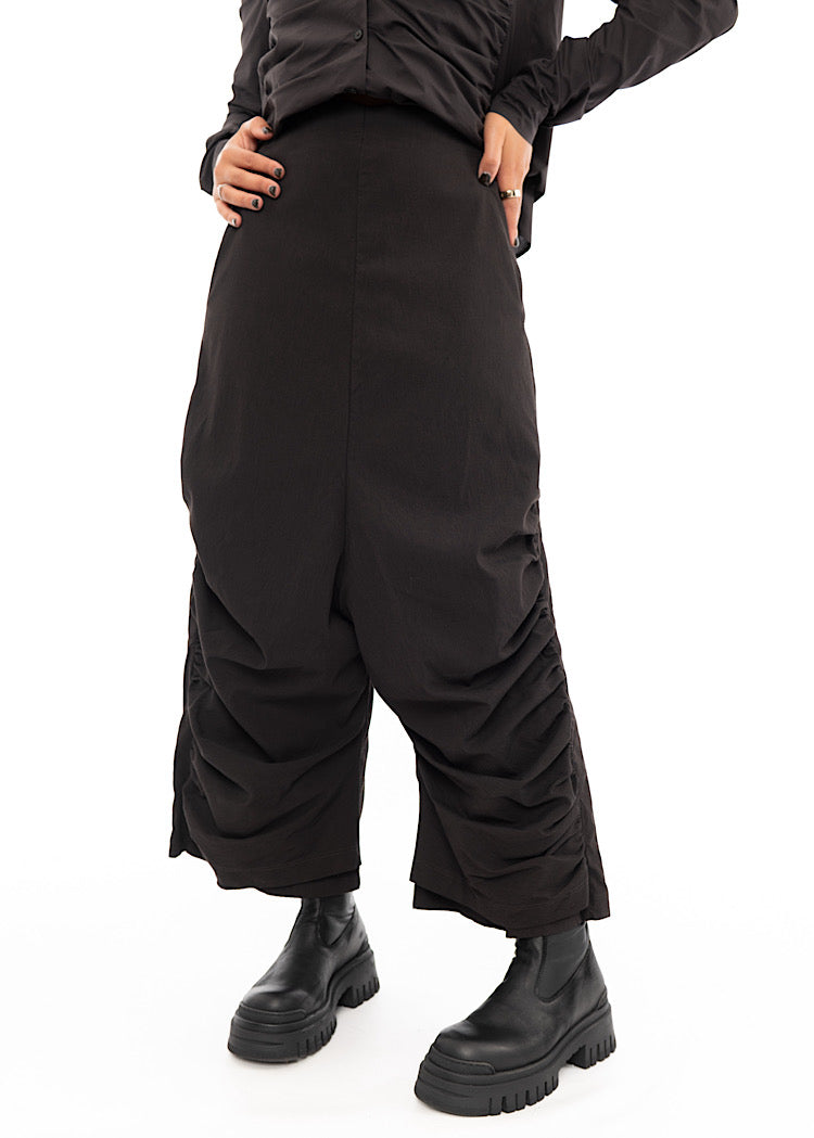 A Model standing slightly to an Angle, wearing a drop-crotch Trouser in a washed dark Brown colour. Showing the straight fit of the waist.