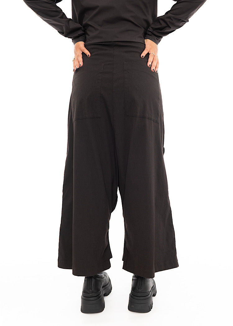 A Model standing facing the back, wearing a drop-crotch Trouser in a washed dark Brown colour. Focusing on the leg shape.