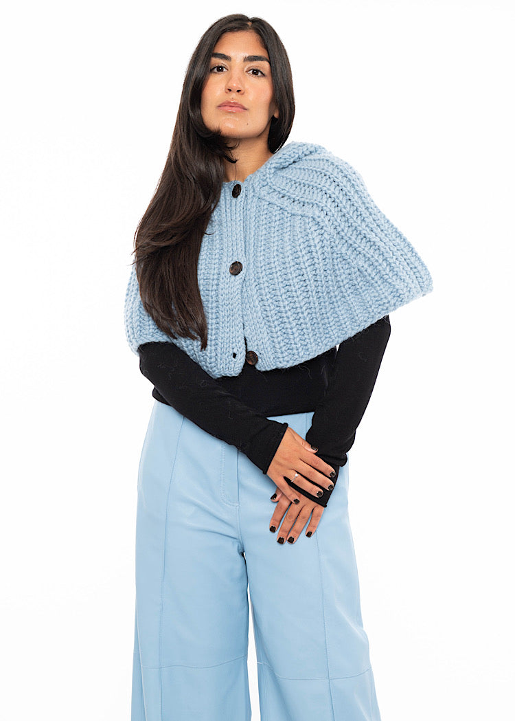 A model wearing an oversized knitted shawl with a hood in light blue. Showing the minimalistic buttons and the length of piece. Styled with a fitted black pullover and light blue leather trousers.