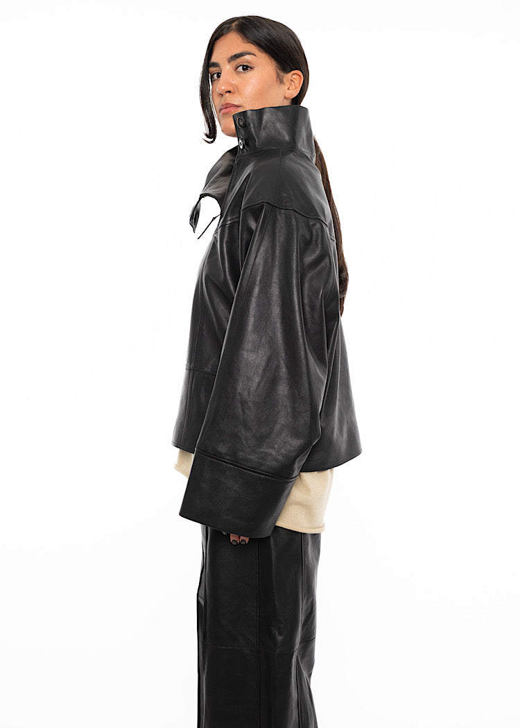 A model standing to a side angle wearing an oversized Black Leather pullover showing the width of the arms and the volume the collar creates.
