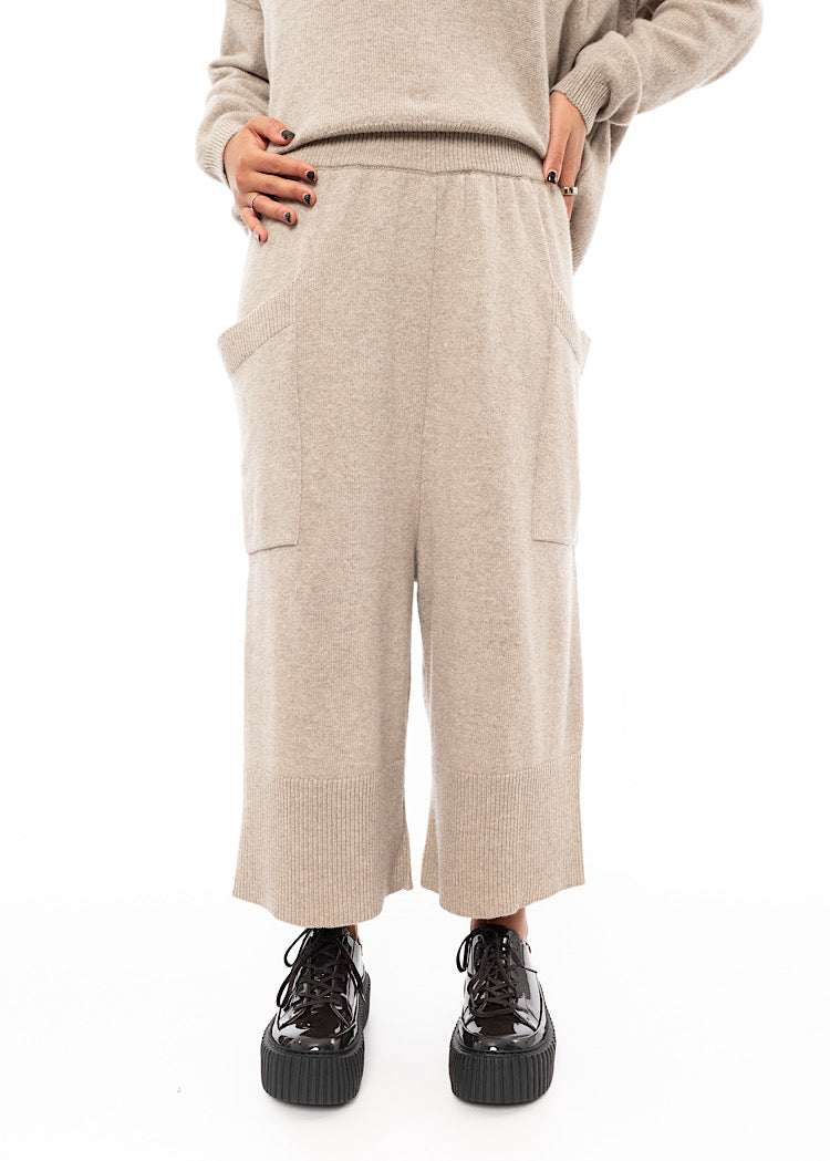 A Model wearing a light beige knitted Trouser. Paired with a matching pullover and Brown patent Shoes.Sowing the straight fit of the piece.