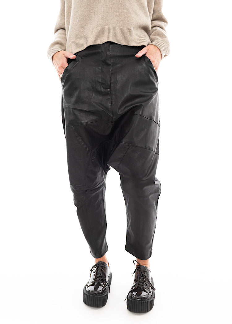 A Model wearing a Structured Drop-Crotch Black Leather Trouser, disclosing the pockets. Styled With a patent Brown Shoe and a creme pullover.