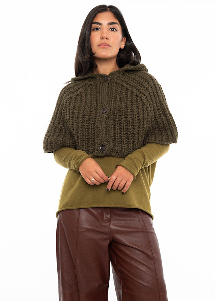 A model wearing an oversized knitted shawl with a hood in dark green. Styled with a fitted khaki green pullover and burgundy leather trousers.