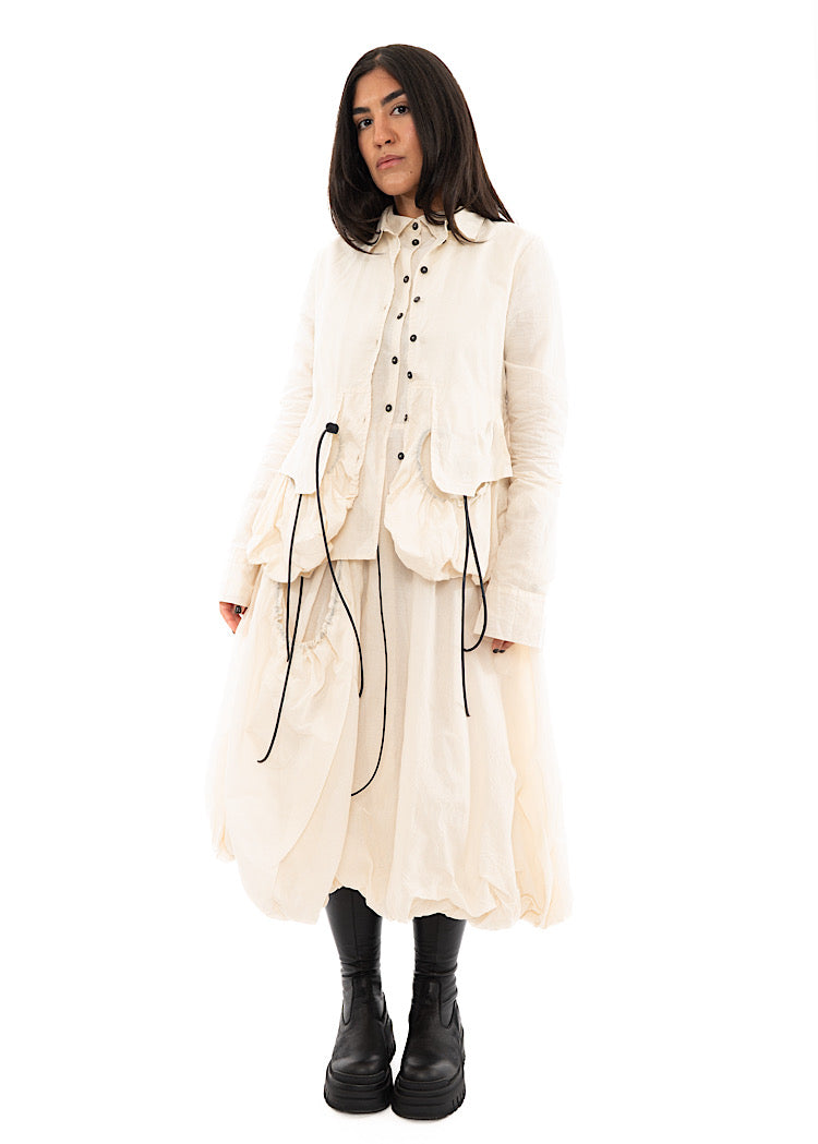 A full frame photo of A Model wearing a bubble skirted Dress in a creme colour. Focusing on the flap pocket, paired with a Black Boot and matching Jacket.