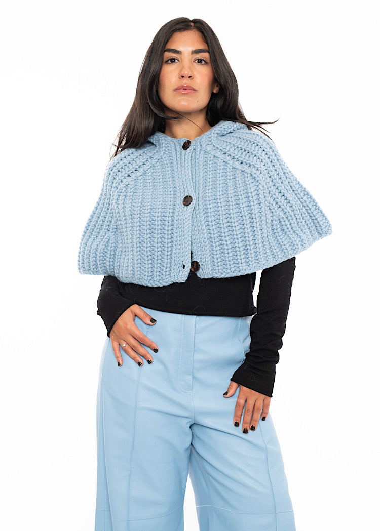 A model wearing an oversized knitted shawl with a hood in light blue. Styled with a fitted black pullover and light blue leather trousers.
