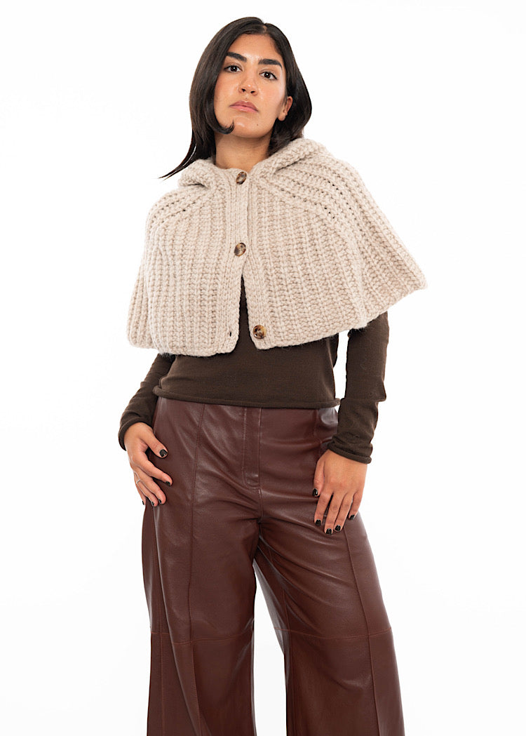 A model wearing an oversized knitted shawl with a hood in creme with beige minimal buttons to fasten. Styled with a fitted black pullover and burgundy leather jeans.