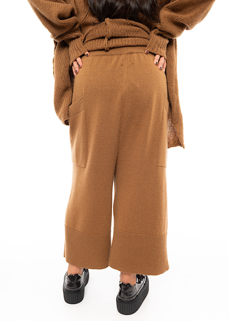 A Model facing the back wearing a Knitted pair of off-orange Trousers, paired with a matching pullover and patent shoes.