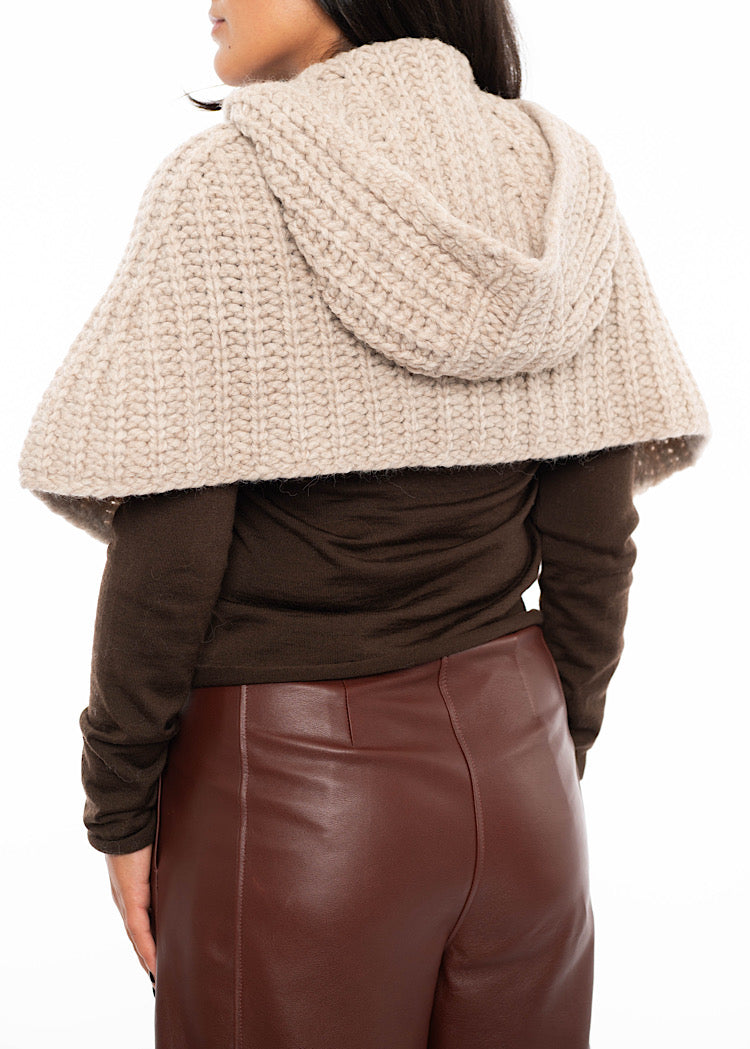 A model standing facing the back wearing an oversized knitted shawl with a hood in creme with beige minimal buttons to fasten. Showing the how the hood sits on the back of piece.