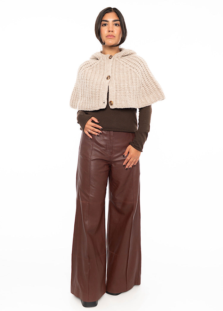 A model wearing an oversized knitted shawl with a hood in light beige with brown buttons to fasten. Styled with a fitted black pullover and burgundy leather jeans.