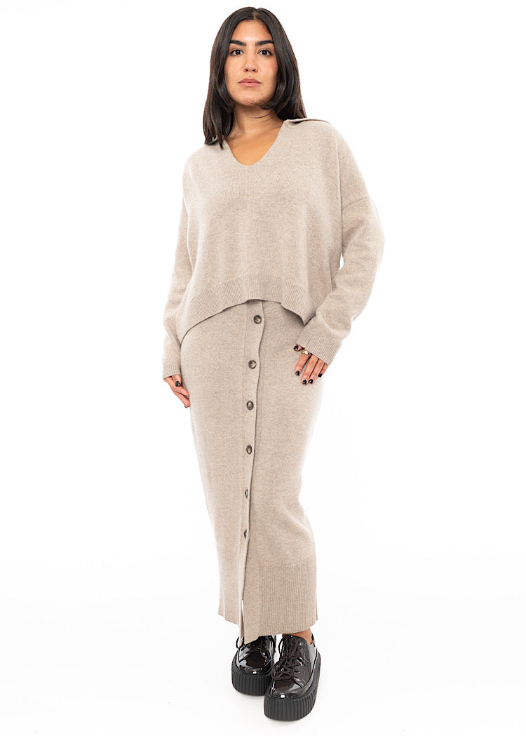 A full frame photo of A Model wearing a light beige completely buttoned up Skirt. Paired with a matching pullover and patent Brown Shoe.