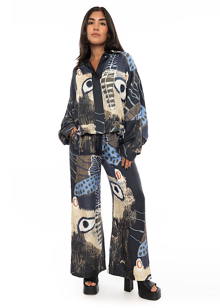 A model wearing an oversized silk shirt with multicoloured print throughout. With hidden buttons to fasten and styled with matching silk wide leg trouser and black high heels.