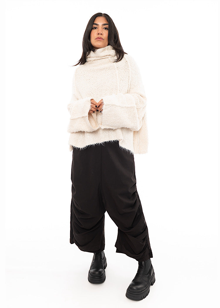 A full size photo of A Model wearing a Light creme, Fluffy pullover featuring a turtle neck feature and an oversized fit. Paired with Black trousers and Black Boot.
