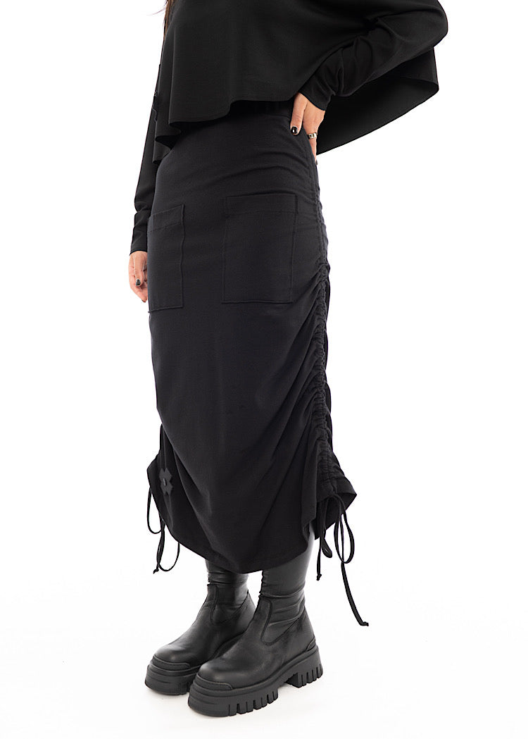 A Model standing slightly to the side wearing a Black drawstring Skirt, styled with a cropped Black Pullover and Chunky Black Boot. Showing the amount of tie on skirt.