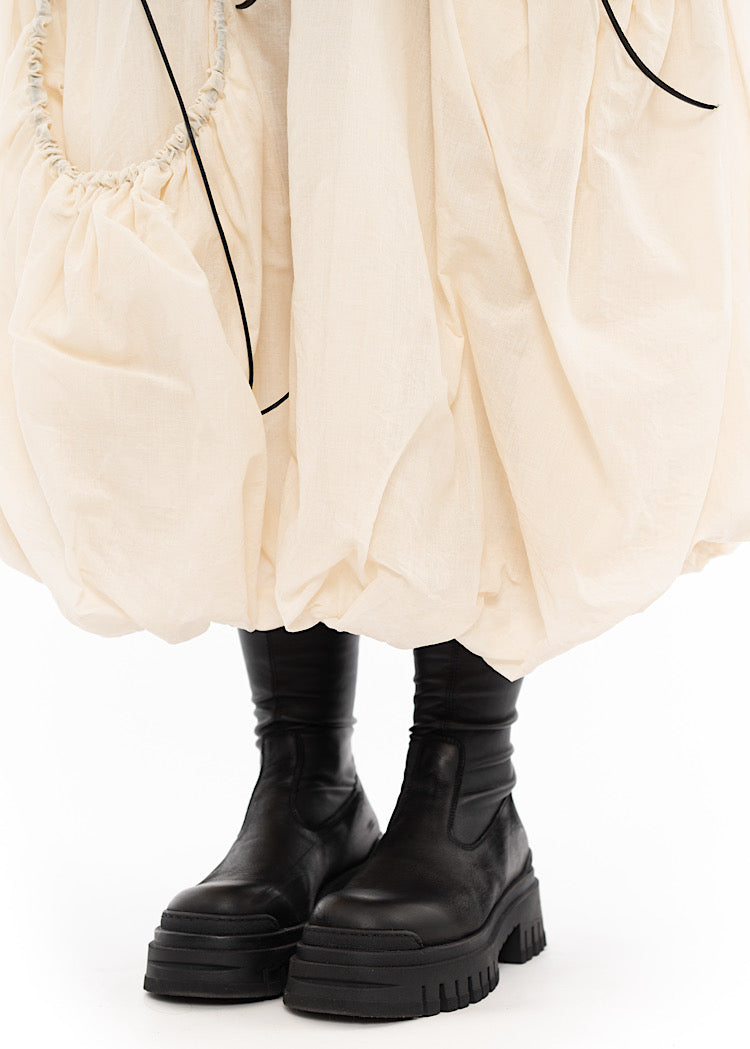 An up close off A Model wearing a bubble skirt in a creme colour. Focusing on the Hem of the Skirt, which is paired with a Chunky soled Boot.
