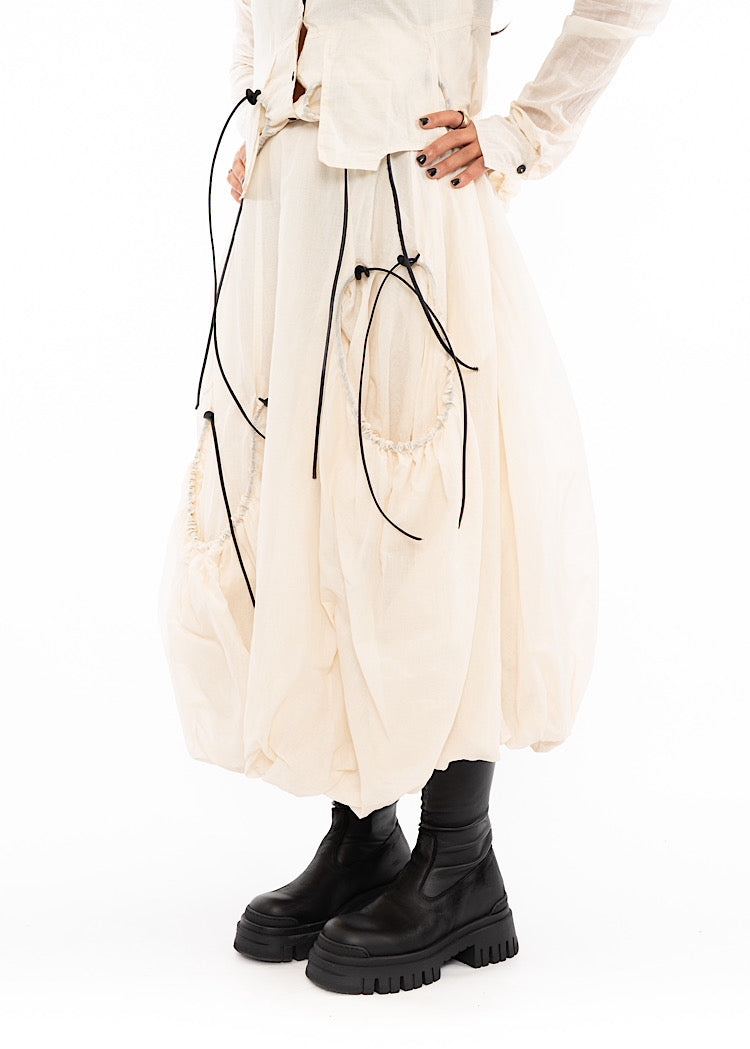 An up close off A Model wearing a quilted bubble skirt and Jacket, in a matching creme colour. Focusing on the Black string details and distinct pockets.