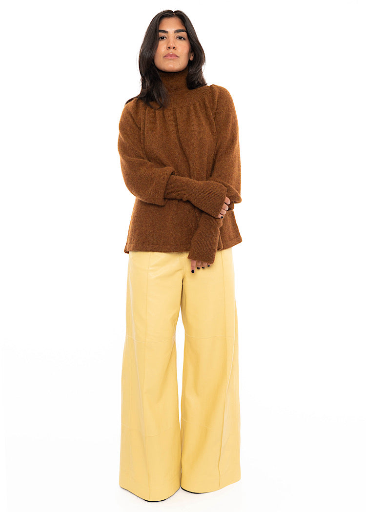 A model wearing a high neck relaxed fit pullover, showing ribbed detailing on the neckline and cuffs. Styled with light yellow straight leg leather trousers and black high heels.
