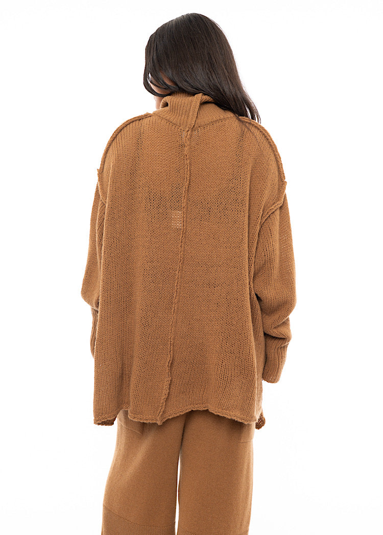 A Model standing facing the back wearing a light brown knitted Oversized Pullover. Showing the long length from the back.