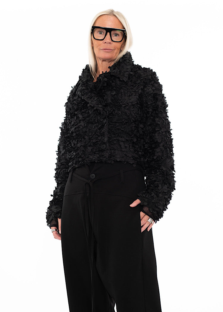 Model wearing black cropped textured jacket with 3D floral applique detail, styled with black drawstring drop-crotch trousers