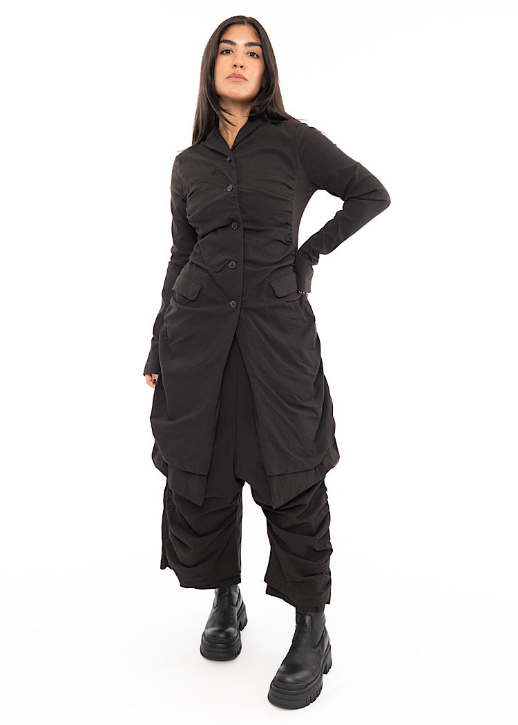 A full frame photo of A Model wearing a fitted techno coat and matching trousers, in a dark off-Brown tone. Paired with a chunky soled Boot.