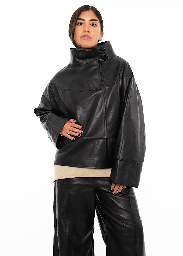 A model wearing an oversized Black Leather pullover with relaxed fit sleeves and a distinctive done up collar. Styled with wide leg trousers in the matching Black leather and black high heels.