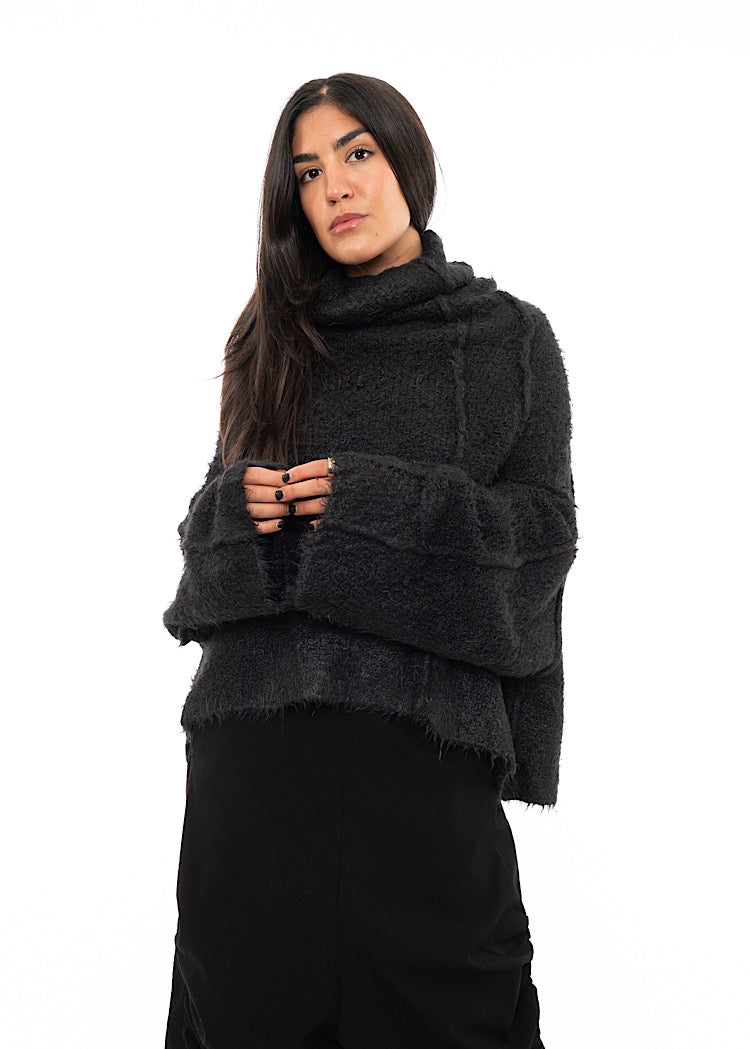 A Model wearing a Dark Grey Pullover, with features of a relaxed turtle neck and wide long sleeves. Showing the oversized relaxed look, paired with a fitted Trouser.