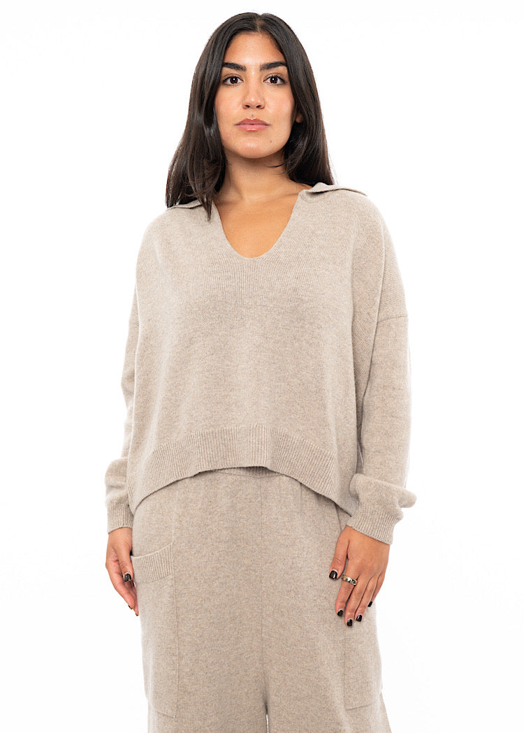 A Model wearing a light Beige knitted pullover with a ribbed hem and a deep V-neckline.Styled with a matching Trouser.