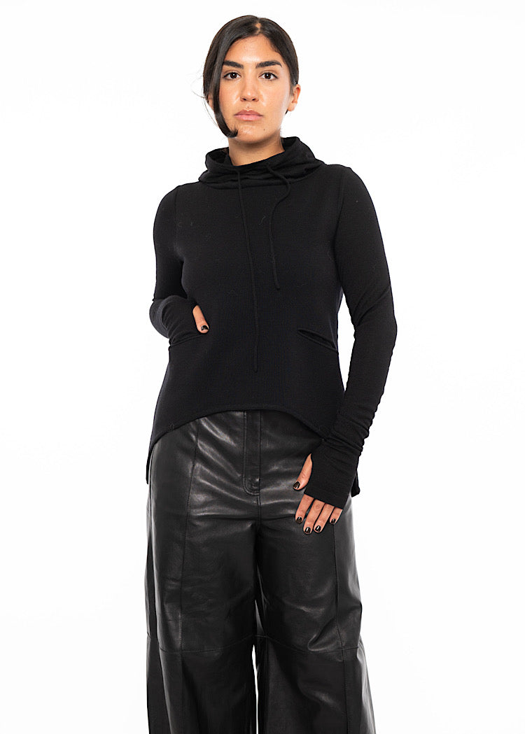 A model wearing a fitted black pullover with a drawstring high neckline, with an asymmetric hemline. Styled with wide leg black leather trousers.