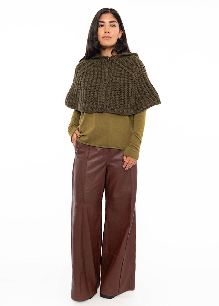 A model wearing an oversized knitted shawl with a hood in dark green. Styled with a fitted khaki green pullover and Burgundy leather trousers and black high heels.