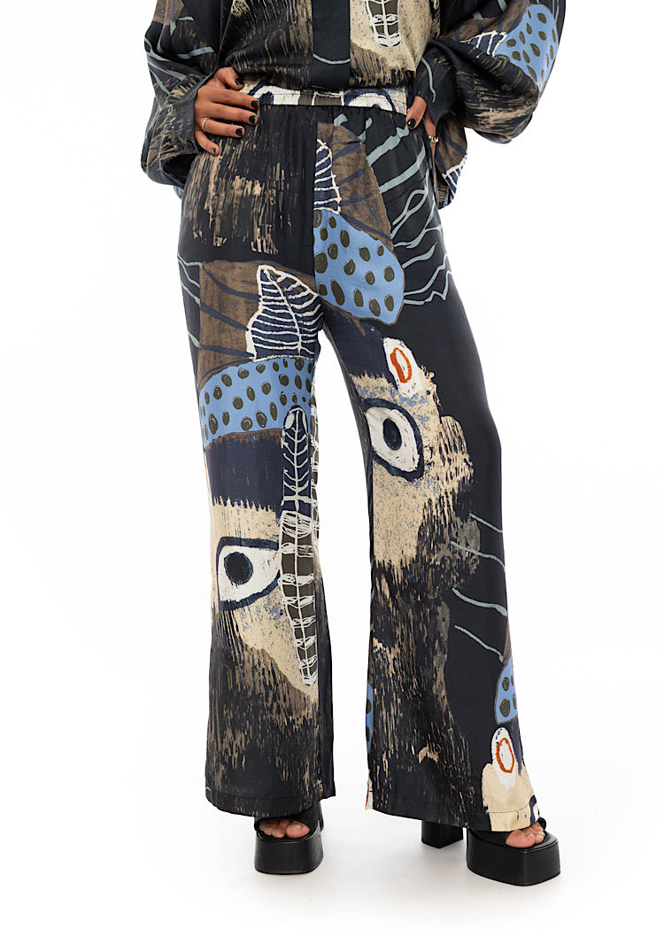 A close up of a model wearing wide leg Silk trousers with a multicoloured pattern throughout. Showing the high waisted length and flare style trouser.