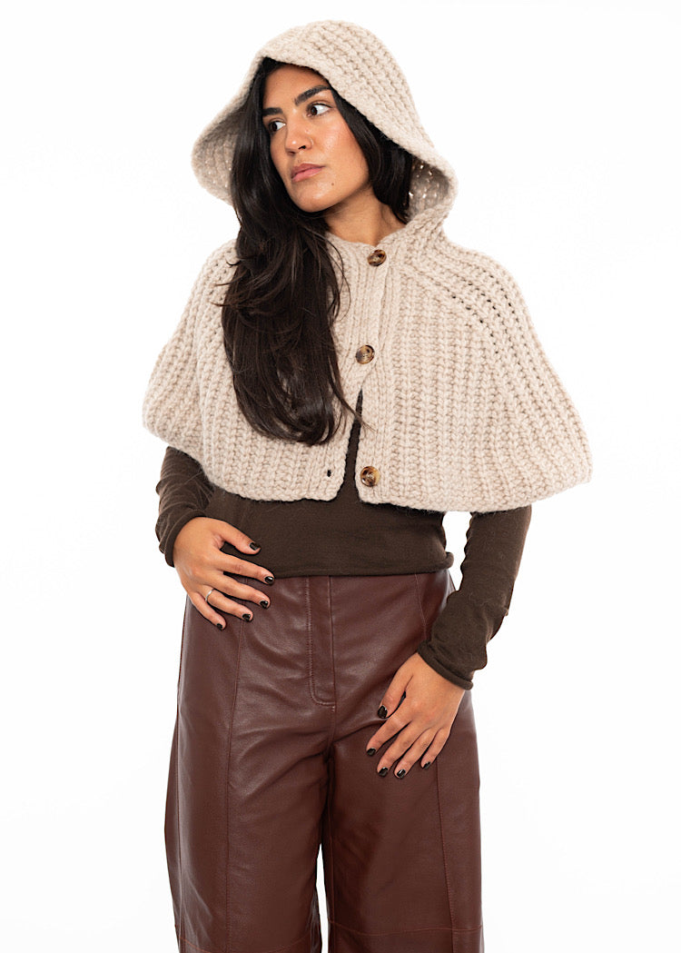 A model wearing an oversized knitted shawl with a hood in creme with beige minimal buttons to fasten. Showing the hoods style up .
