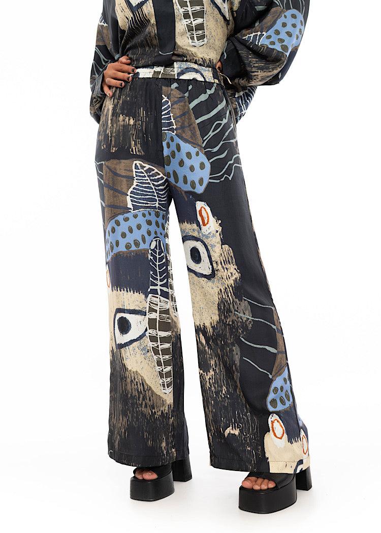 A close up of a model wearing a wide leg Silk trousers with a multicoloured design throughout. Showing the high waisted length and floaty style trouser.