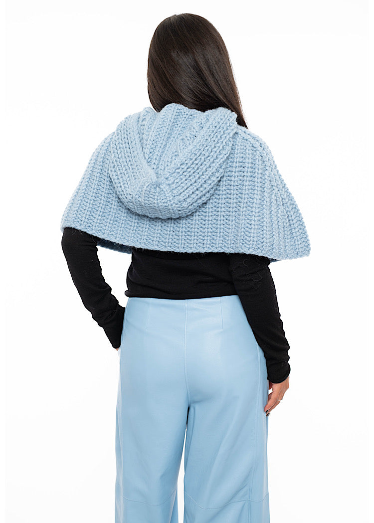A model standing facing the back wearing an oversized knitted shawl in light blue. Showing the back length and hood in more detail.