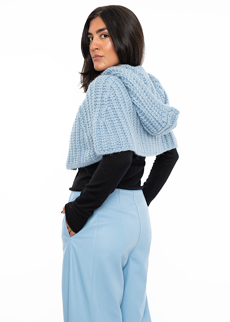A model standing to a side angle wearing an oversized knitted shawl in light blue. Showing the width and hood in more detail.