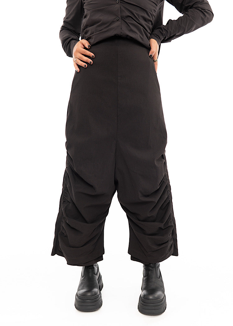 A Model wearing a drop-crotch Trouser in a washed dark Brown colour. With a kick-flare leg, paired with a matching straight fit shirt.