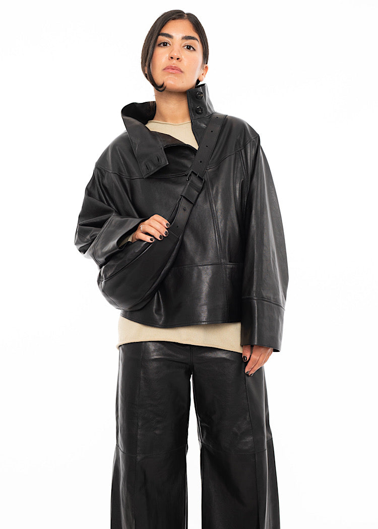 Close up of Model wearing a black Leather jacket, with a matching trouser. Holding Black Slouchy leather bag on a longer strap which is hanging over models shoulder disclosing buckle detail.