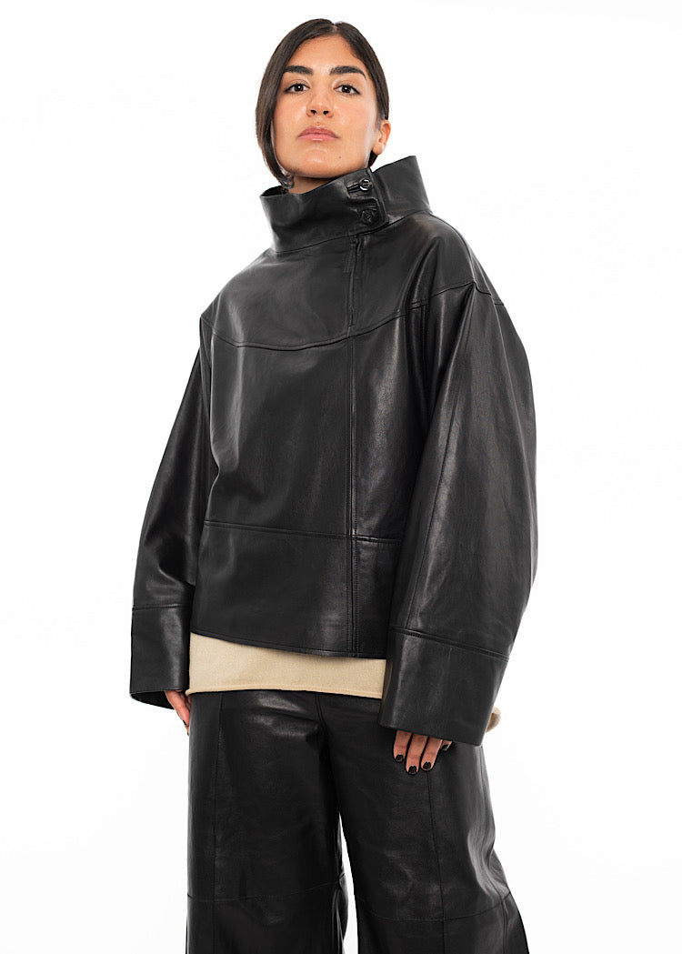A model wearing an oversized Black Leather pullover with relaxed fit sleeves, black zipper detail and a unique done up collar. Styled with matching wide leg trousers.