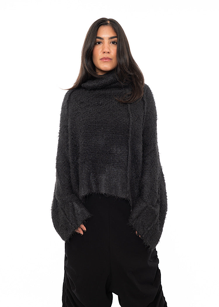 A Model wearing a washed Grey Pullover, including features of a relaxed turtle neck and wide long sleeves. Paired with Black Trousers.