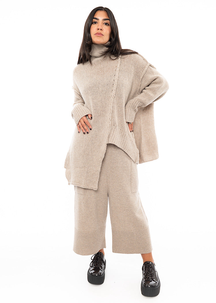 A full frame photo of A Model wearing a creme Knitted Pullover with Distressed pieces throughout. Showing the seam-lines in more detail.