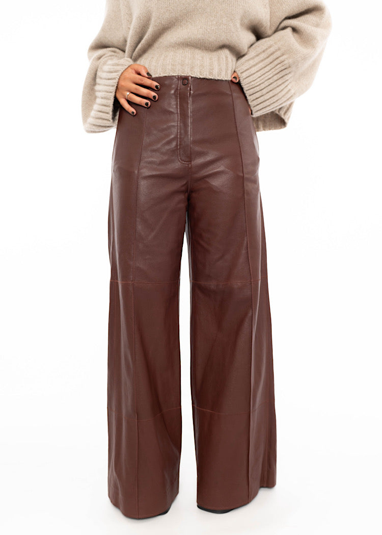 Model wearing Burgundy wide leg leather trousers with a zipper and button to fasten. Styled with an oversized warm beige pullover with ribbed detailing.