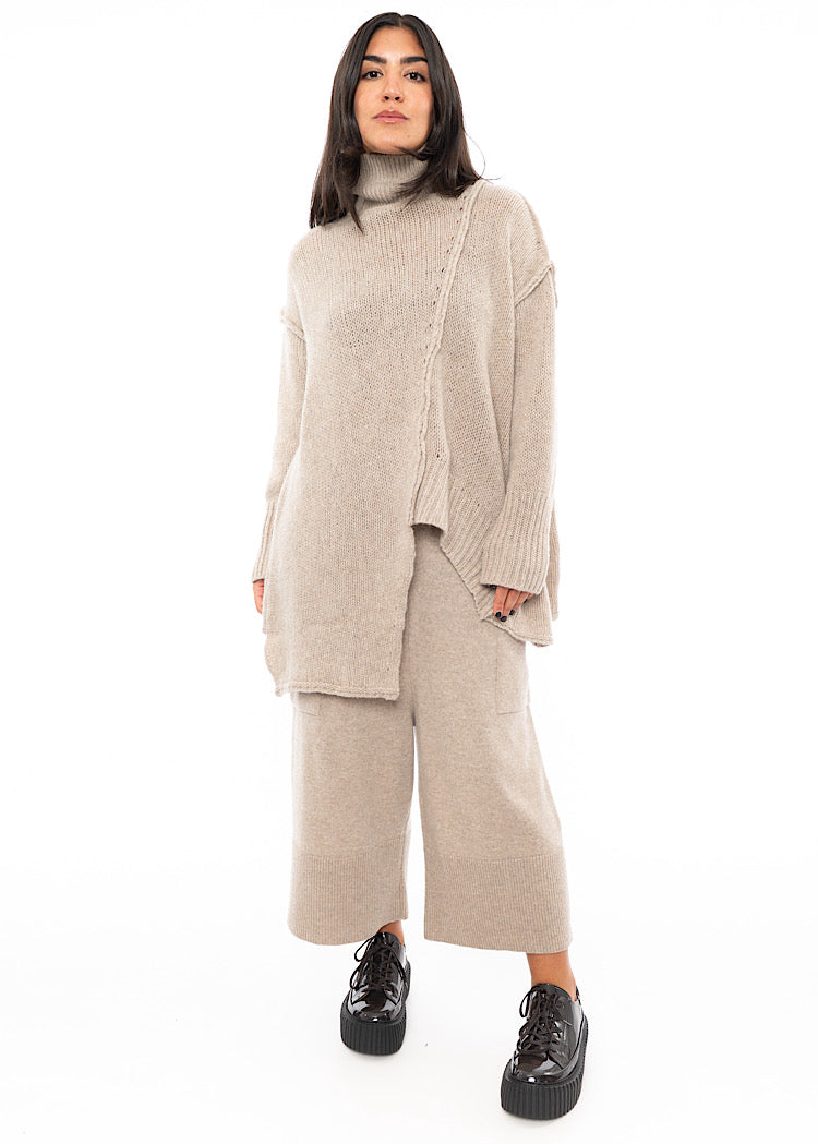 A full frame photo of A Model wearing a creme Knitted Pullover with Distressed pieces throughout. Paired with a matching Trouser and patent Shoe.