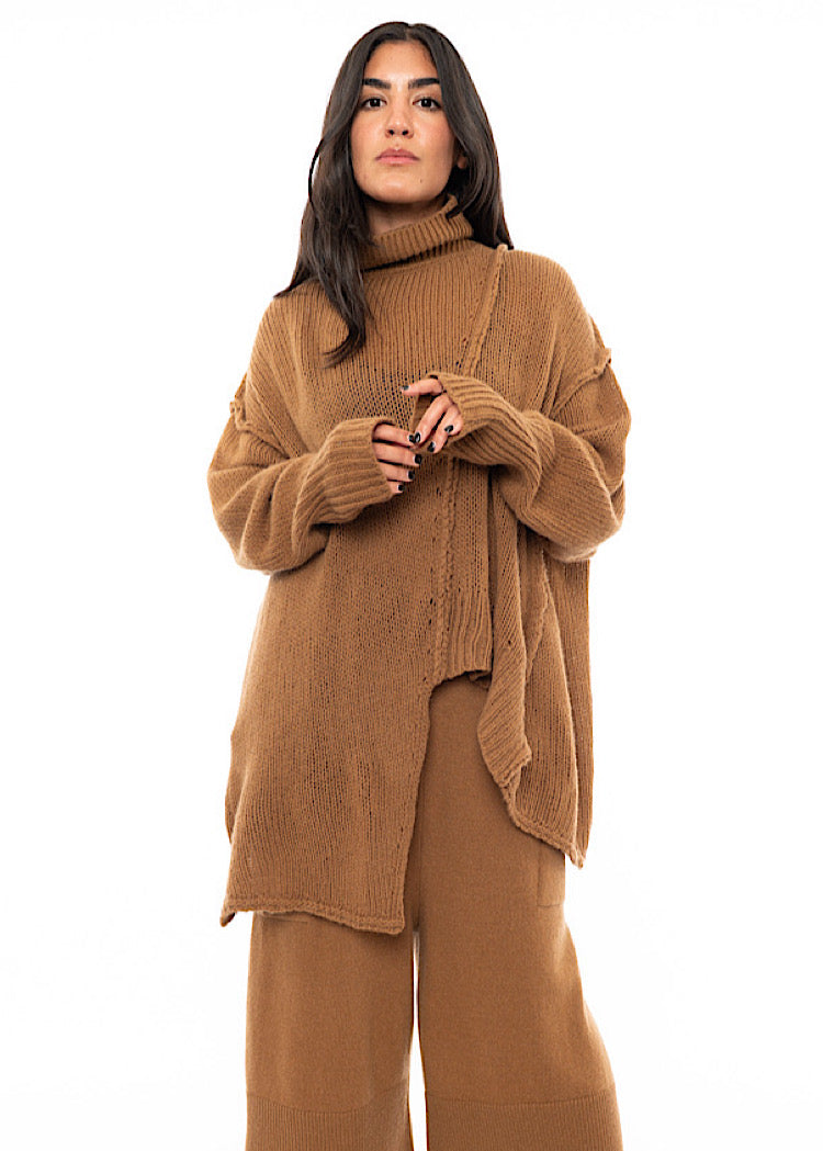 A Model wearing an off orange knitted Oversized Pullover with distressed pieces dangling down. Paired with a matching tone Trouser.