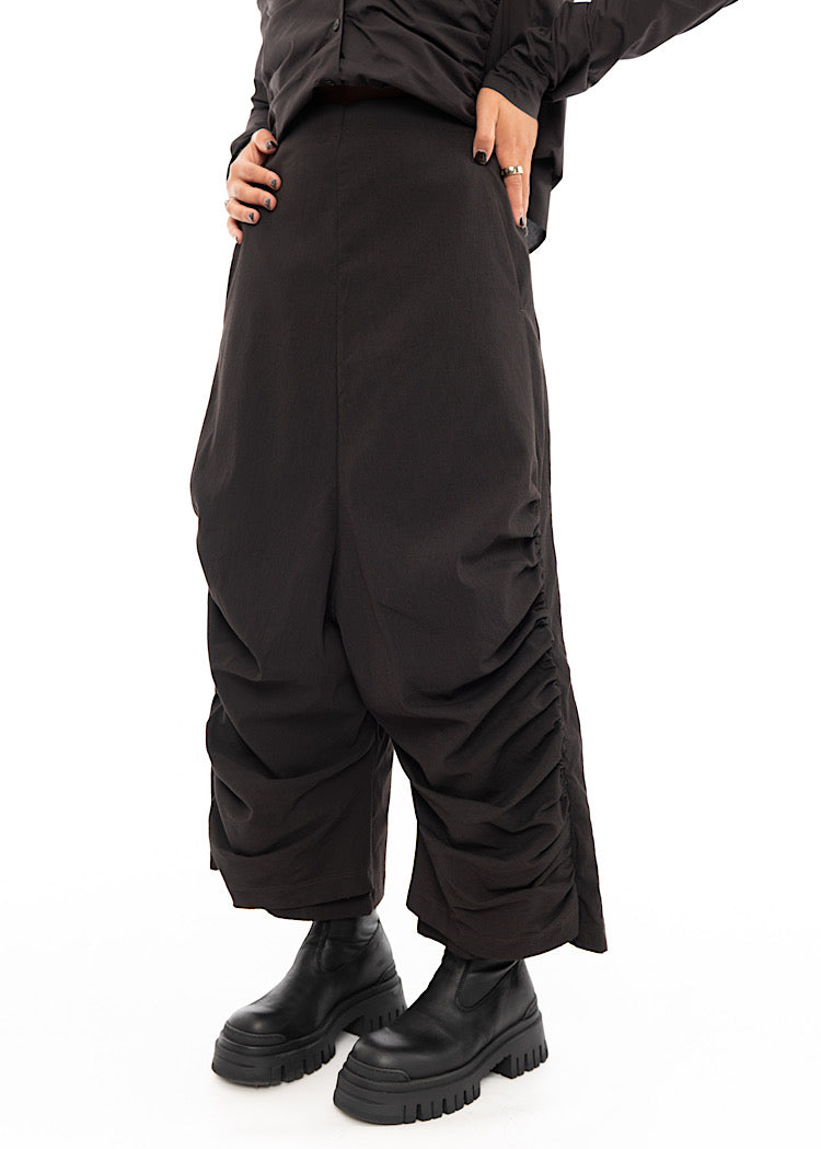 A Model standing slightly to an Angle, wearing a drop-crotch Trouser in a washed dark Brown colour. Showing the straight fit of the waist.