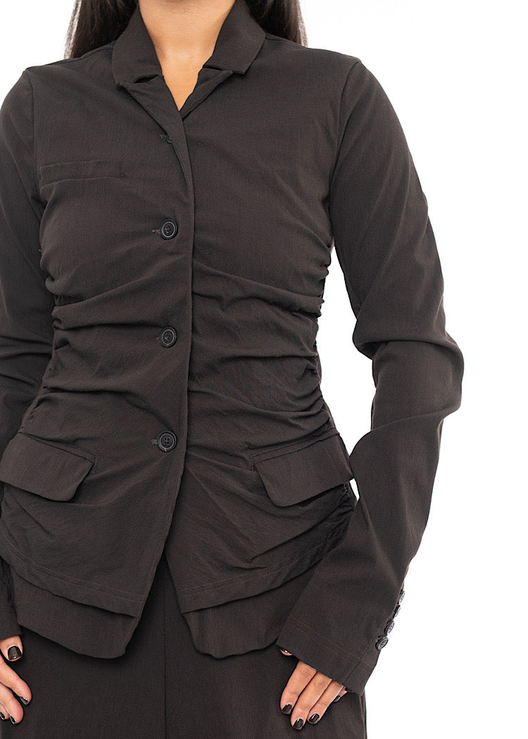 An up close of A Model standing to the side wearing a detailed techno Jacket and matching trousers, in a dark off-Brown colour. Showing the fitted design on the piece.