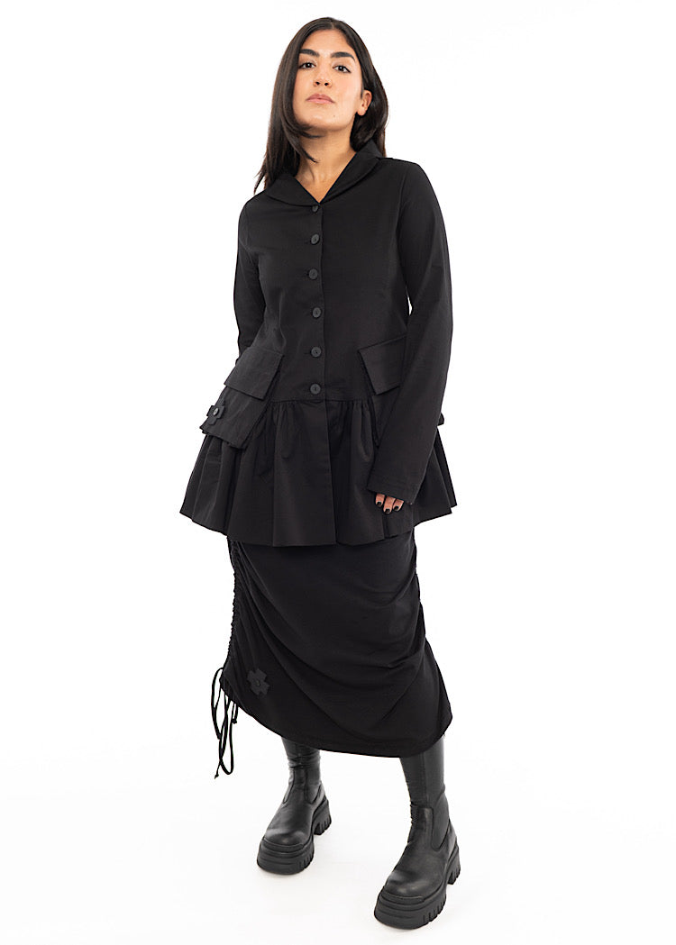 A Model wearing a Black relaxed fit Cotton Jacket, featuring long sleeves, a collared necklines and buttons to fasten. Styled with a matching drawstring skirt and Black Boot.