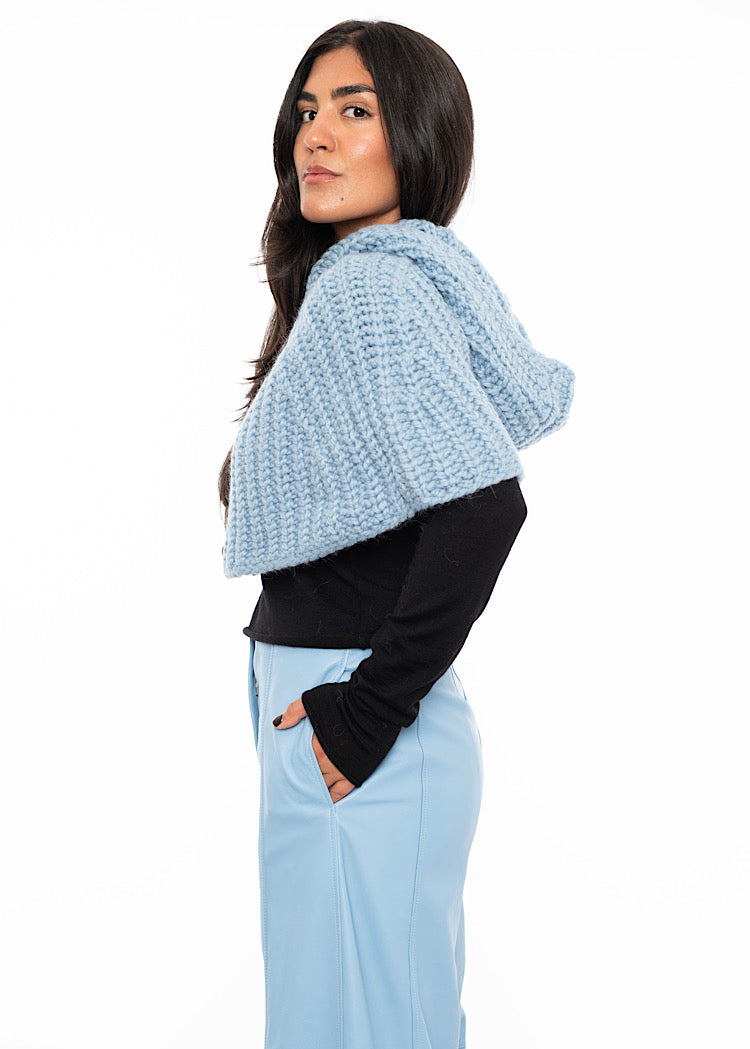 A model standing to a side angle wearing an oversized knitted shawl in light blue. Showing the width and hood in more detail.Styled with a fitted black pullover and light blue leather trousers.