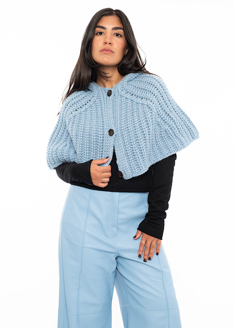 A model wearing an oversized knitted shawl with a hood in light blue. Showing the minimalistic buttons. Styled with a fitted black pullover and light blue leather trousers.
