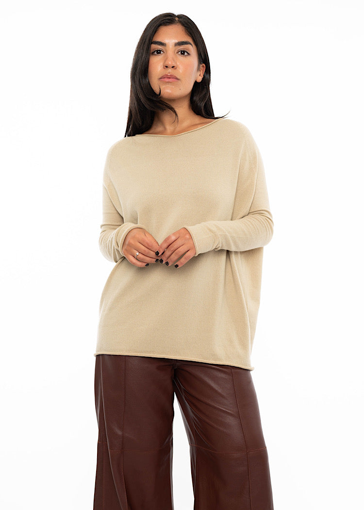A Model wearing a relaxed fit beige pullover with a slash neckline and long sleeves, finishing at the hip. Styled with burgundy wide leg leather trousers. Showing the gathering on the arms.