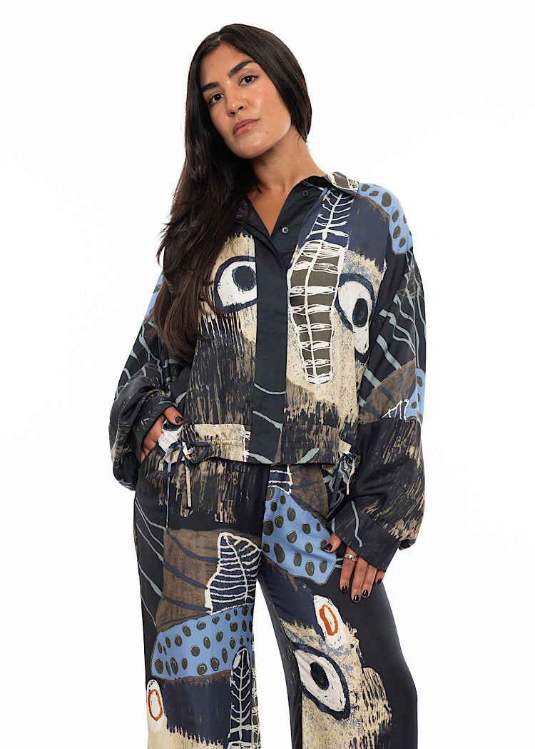 A model wearing an oversized silk shirt with a patterned print throughout. Showing the floaty cuff detailing on the arms, with a gathering effect.Paired with a matching silk wide leg trouser to add volume paired with black high heels.