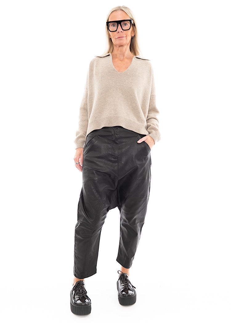 A full frame photo of A Model wearing a Structured Drop-Crotch Black Leather Trouser, disclosing the pockets. Styled With a patent Brown Shoe and a creme