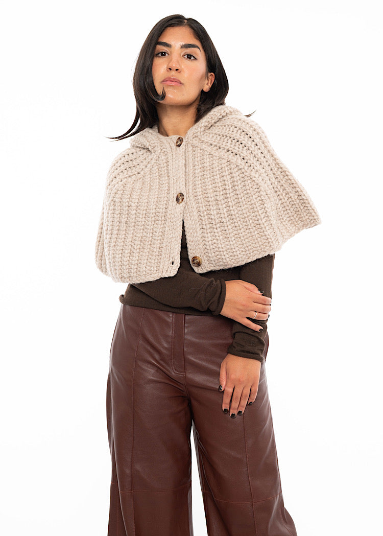 A model wearing an oversized knitted shawl with a hood in creme with beige minimal buttons to fasten. Styled with a fitted black pullover and burgundy leather jeans. Showing the length of shoulders.